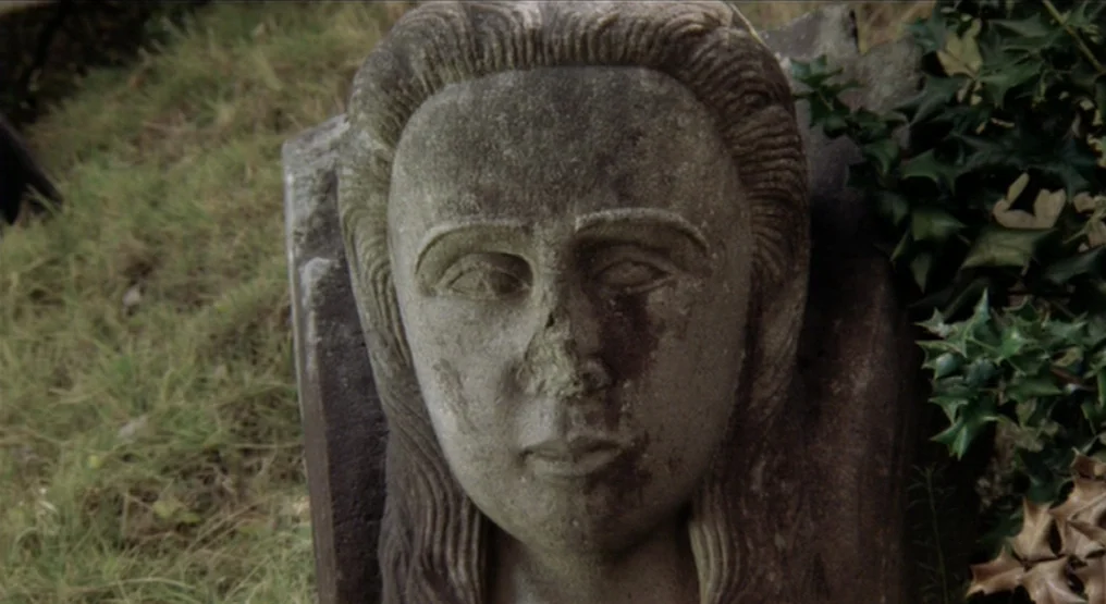 Rhapsody in August (1991) movie still: closeup — A weathered stone bust rests in the grass near a patch of holly. The stone face shows signs of…