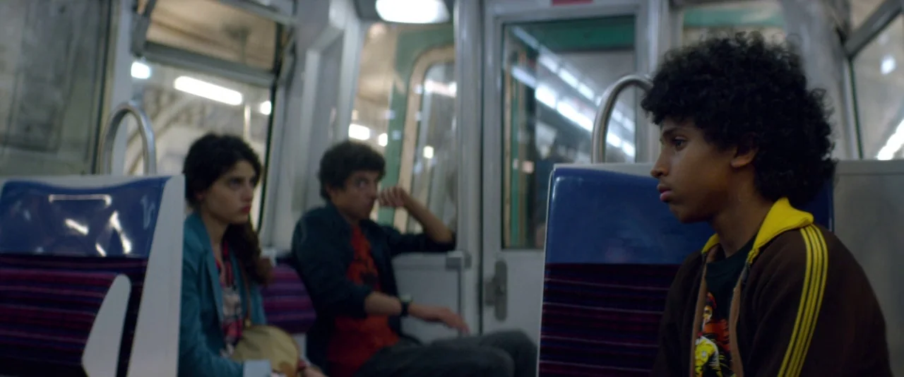 Nocturama (2016) movie still: medium — An interior shot of a public transit car with purple-striped blue seats. Three teenagers occupy the cabin…