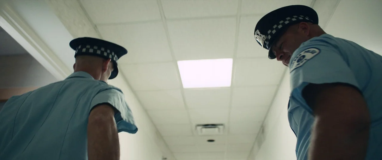 Judas and the Black Messiah (2021) movie still: medium — A long institutional hallway with white walls and a drop-ceiling grid of lights. Two police…