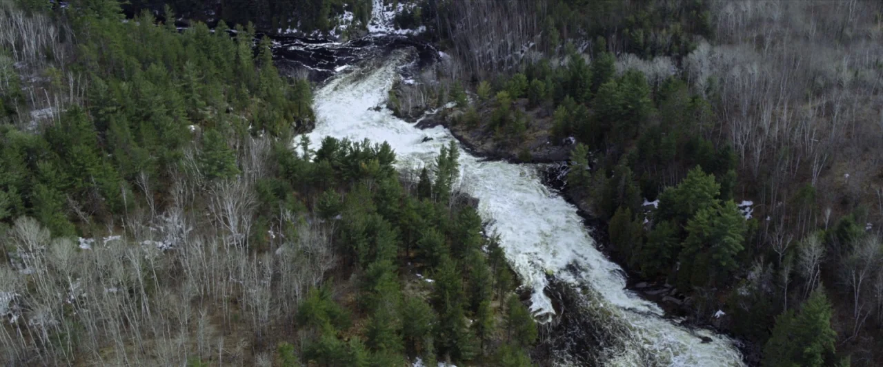 The Silencing (2020) movie still: establishing — A rushing river cuts through a dense forest with patches of snow on ground. White water cascades…