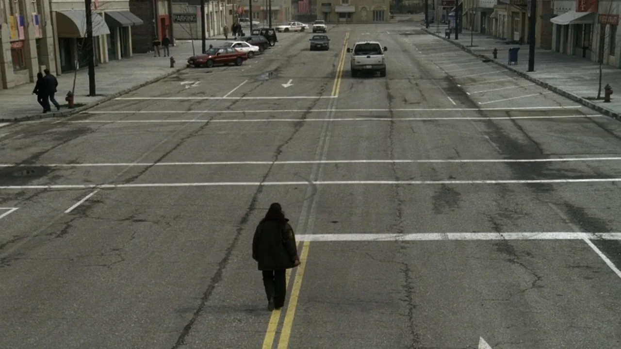 The Shepherd (2023) movie still: wide — An empty asphalt road stretches through a quiet town with storefronts on both sides. A lone individual walks…