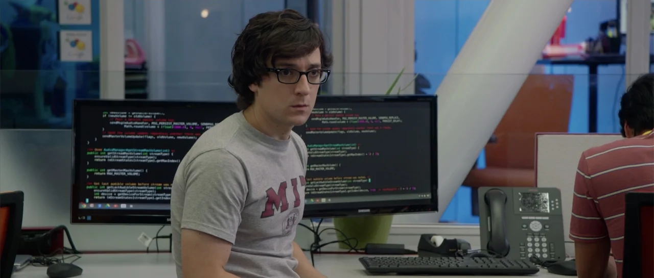 The Internship (2013) movie still: medium — A modern workspace contains desks, computer monitors displaying code, and a standard desk phone. A young…