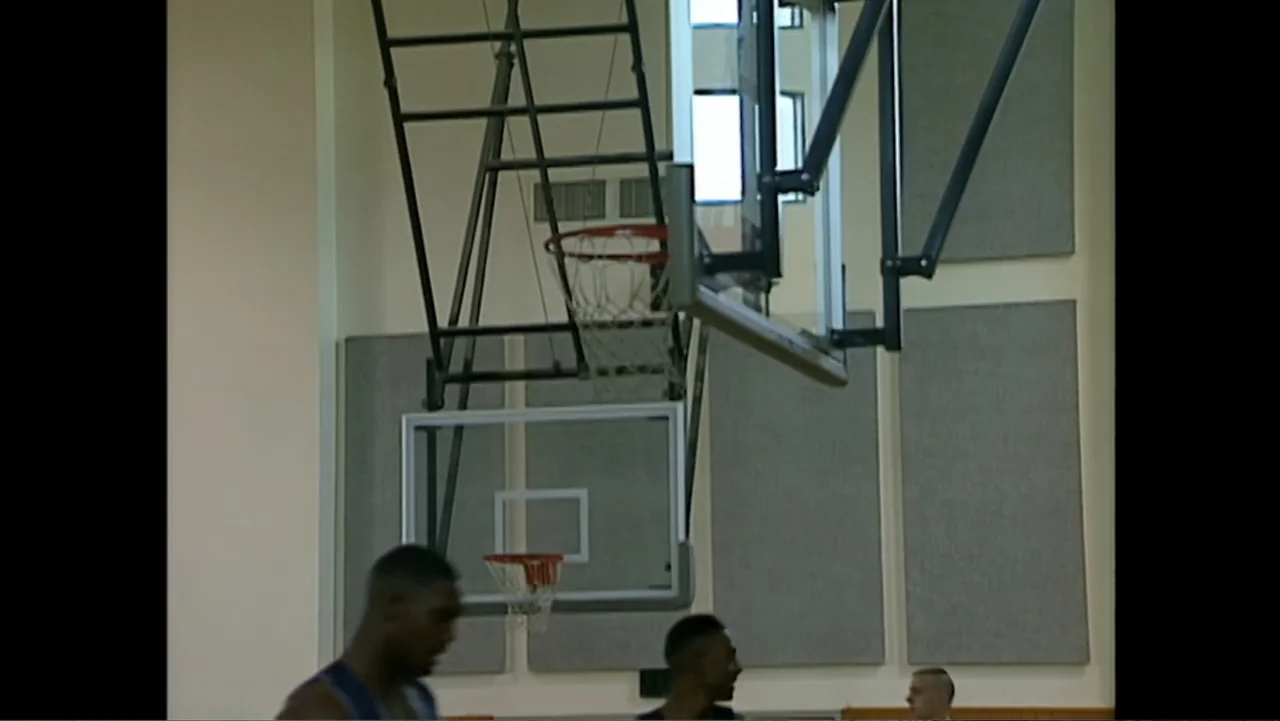 We Beat the Dream Team (2025) movie still: medium — An indoor gymnasium features light walls with grey acoustic panels and a basketball hoop. Three…