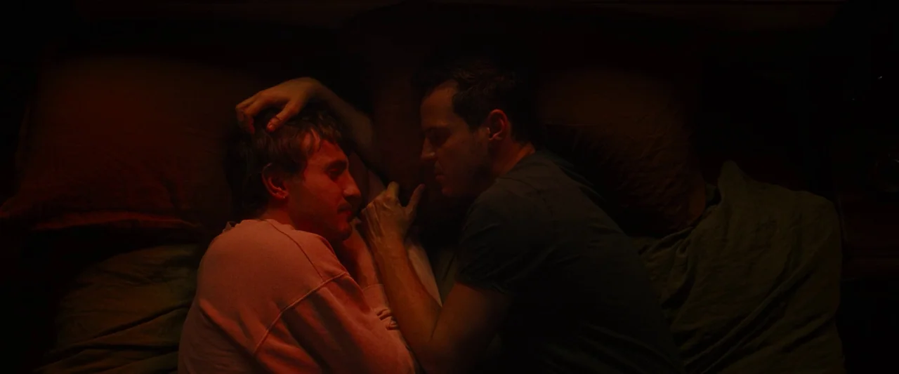 All Of Us Strangers (2023) movie still: medium — Two men lie in a dark setting, close together on a textured surface. The subjects face each other…