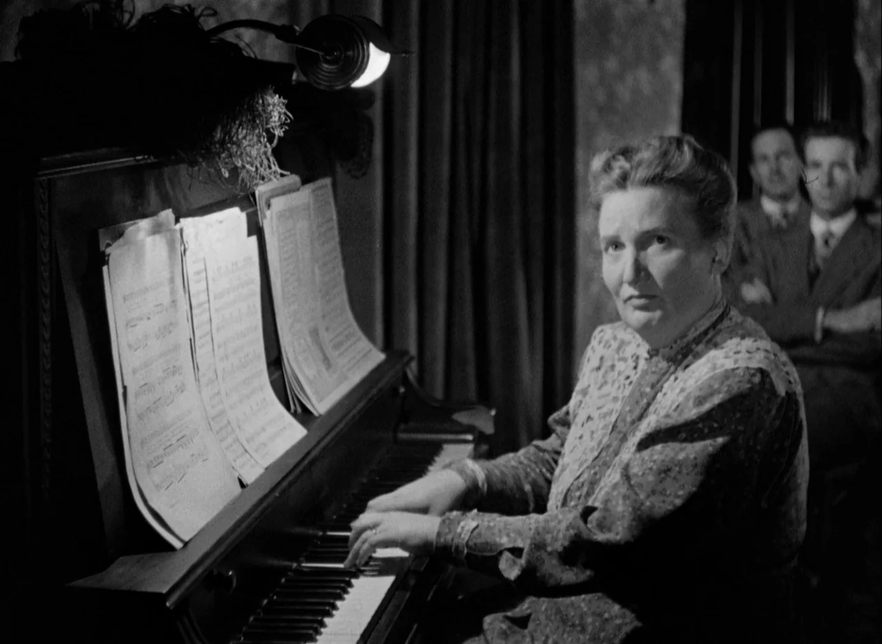 The Spiral Staircase (1946) movie still: medium — A woman plays an upright piano in a dimly lit room with heavy curtains. Two men stand in the…