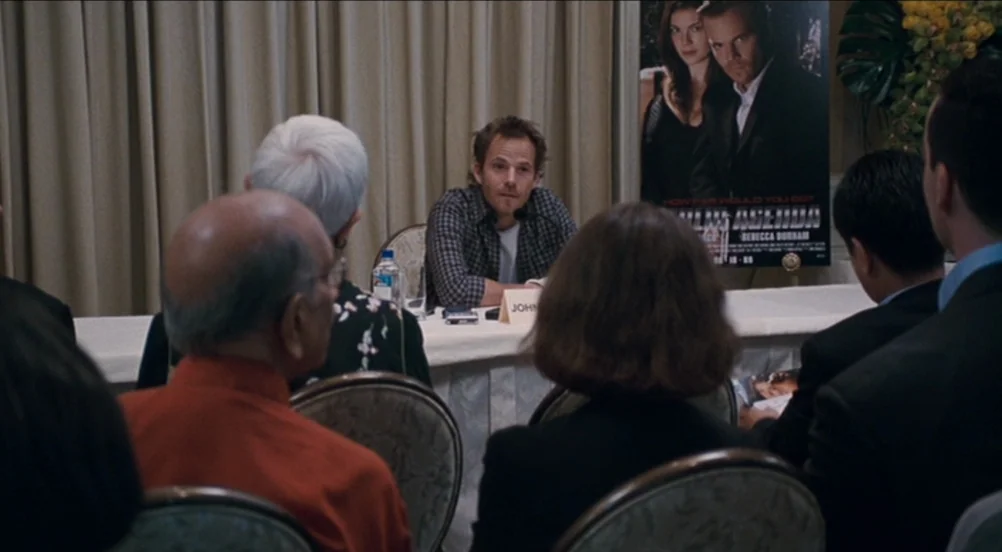 Somewhere (2010) movie still: medium — A man sits alone at a white-clothed table in a room with beige curtains. He is facing an audience of several…