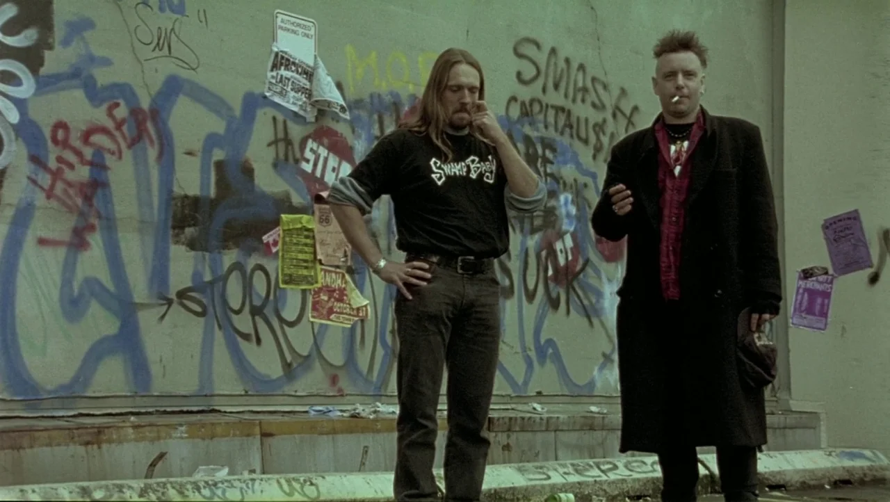 Hard Core Logo movie still: fullbody — Two men stand before a concrete wall heavily covered in layered spray paint graffiti. They occupy a narrow…