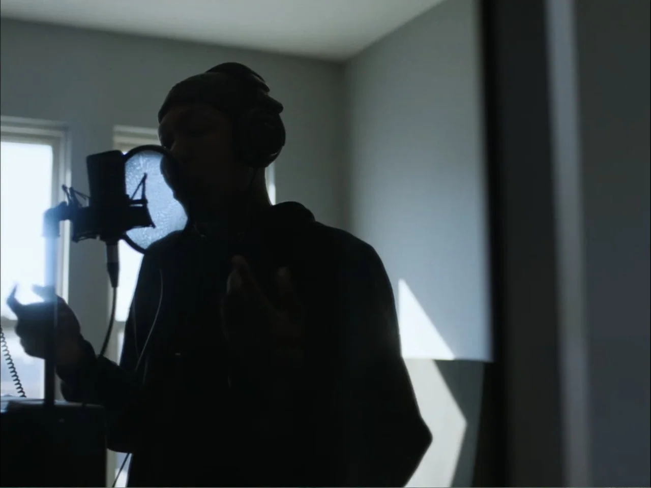 As We Speak: Rap Music on Trial (2024) movie still: medium — A recording space with plain walls and a window providing bright backlighting. A person…