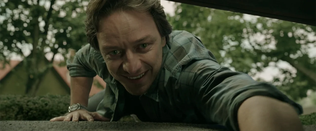 K.G.F: Chapter 2 (2022) movie still: closeup — A man peers over a low concrete wall in an outdoor suburban environment. The man looks down toward…