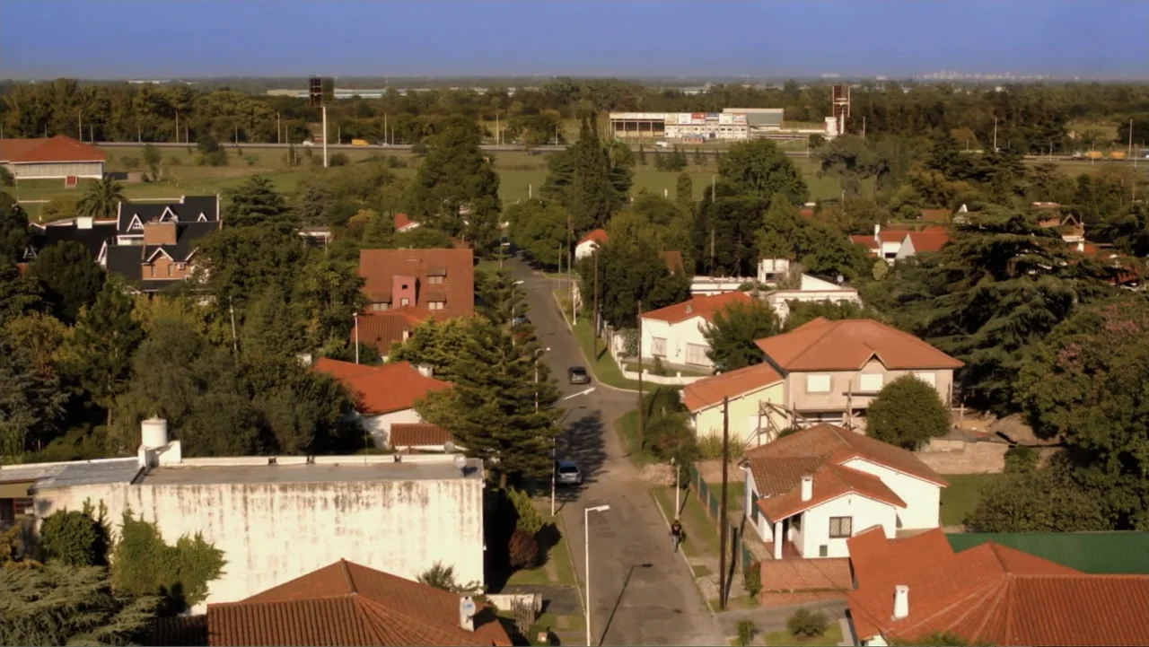 A Boyfriend for My Wife (2008) movie still: establishing — A suburban neighborhood with red-roofed houses sits under a clear blue sky. A paved…