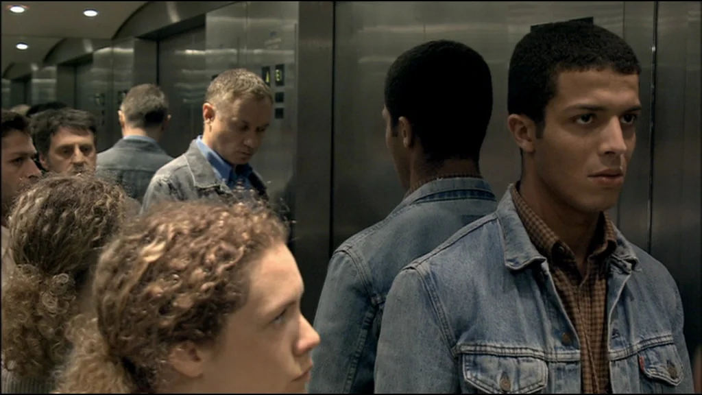 Caché (Hidden) (2005) movie still: medium — A crowded, metallic-walled elevator interior with multiple passengers standing in silence. A young man…