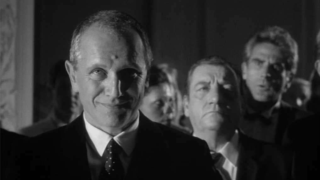 Under The Cherry Moon (1986) movie still: medium — A group of men in suits stand in a dimly lit, crowded indoor space. A man in the foreground…