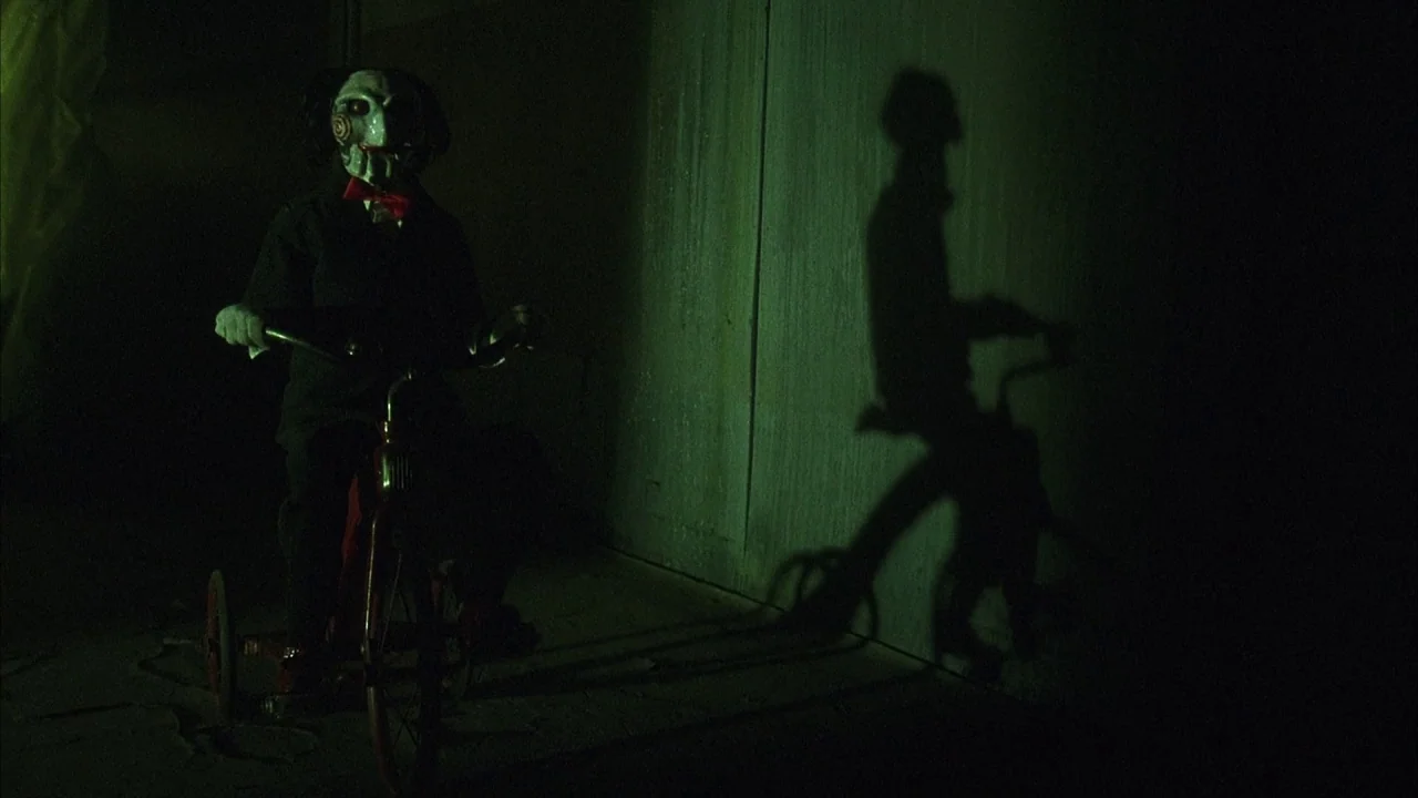 Saw movie still: medium — A dark space features a ventriloquist dummy seated on a small red metal tricycle. The figure casts a large, distorted…