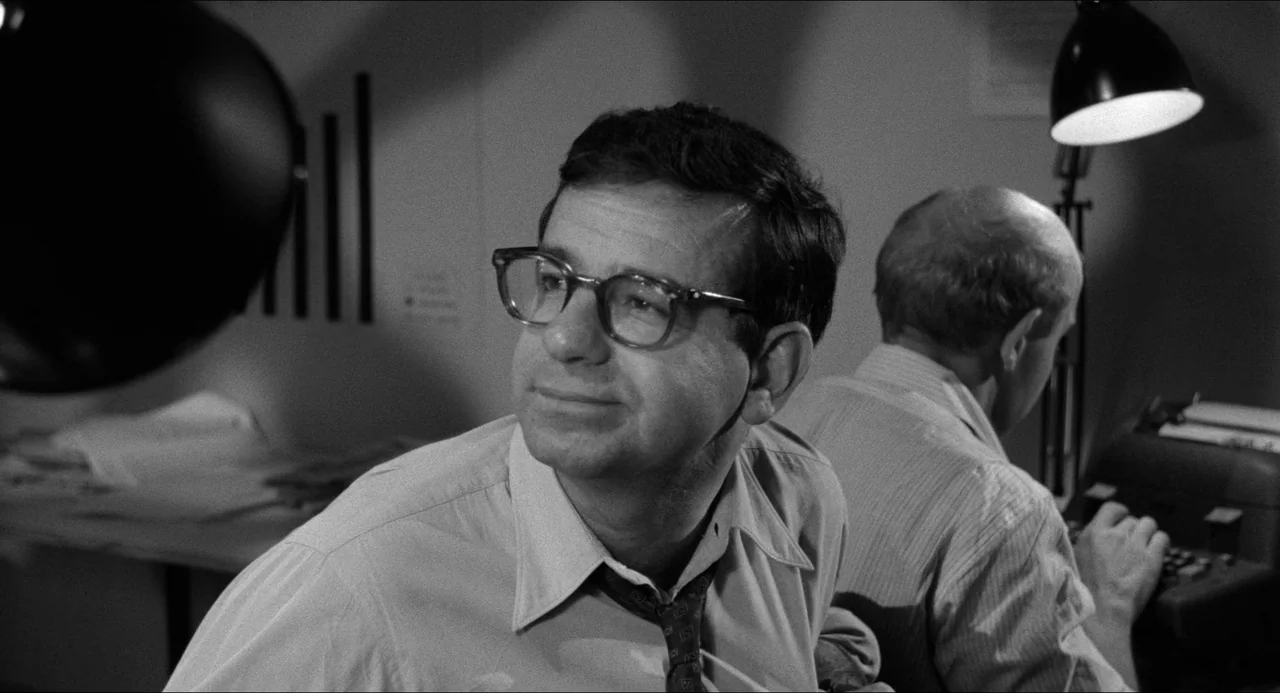 A Face In The Crowd (1957) movie still: medium — A dimly lit room serves as a workspace for two men at their desks. One man types on a machine while…