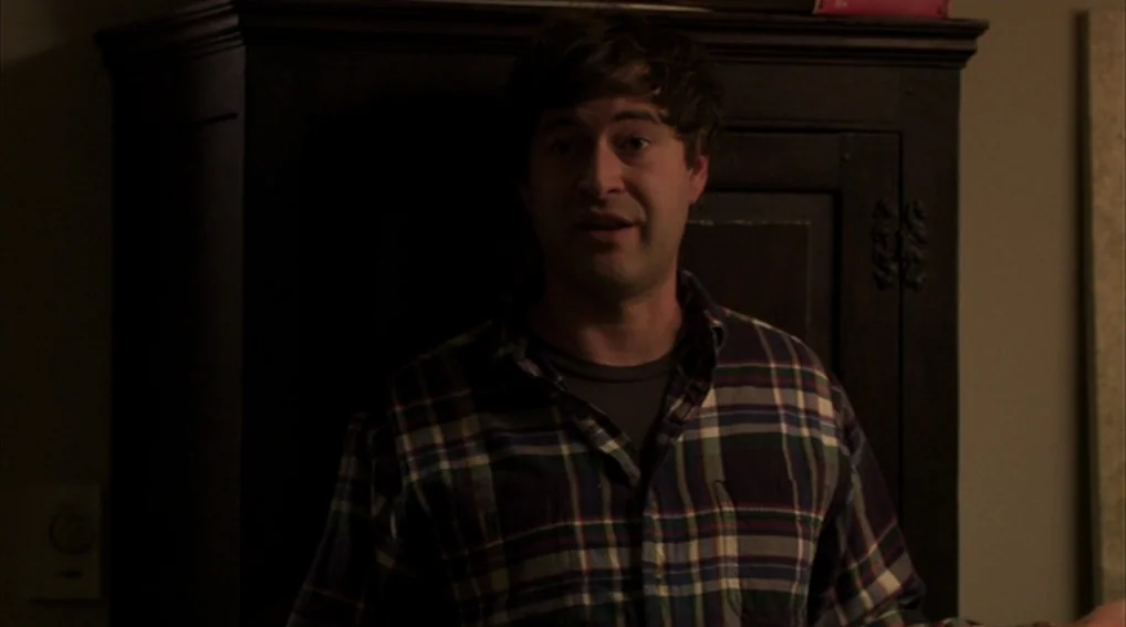 Your Sister’s Sister (2011) movie still: medium — A man stands in a dimly lit room in front of a dark cabinet. He speaks while facing toward the…