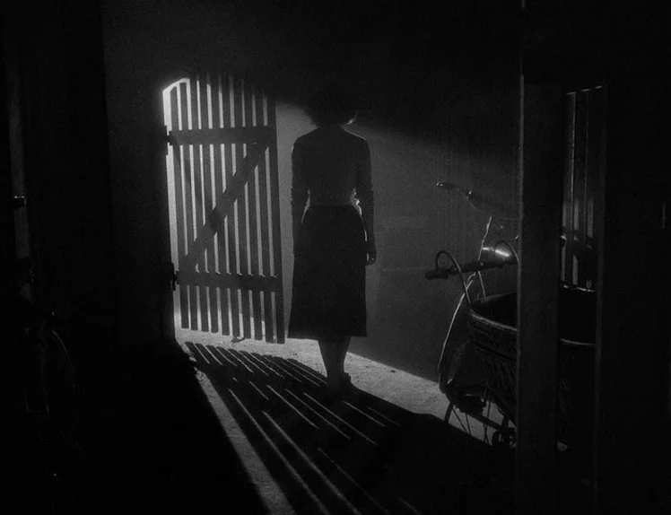 Prison (1949) movie still: fullbody — A dark, wood-paneled interior space with a bicycle parked to the right. A woman stands in the center of an…