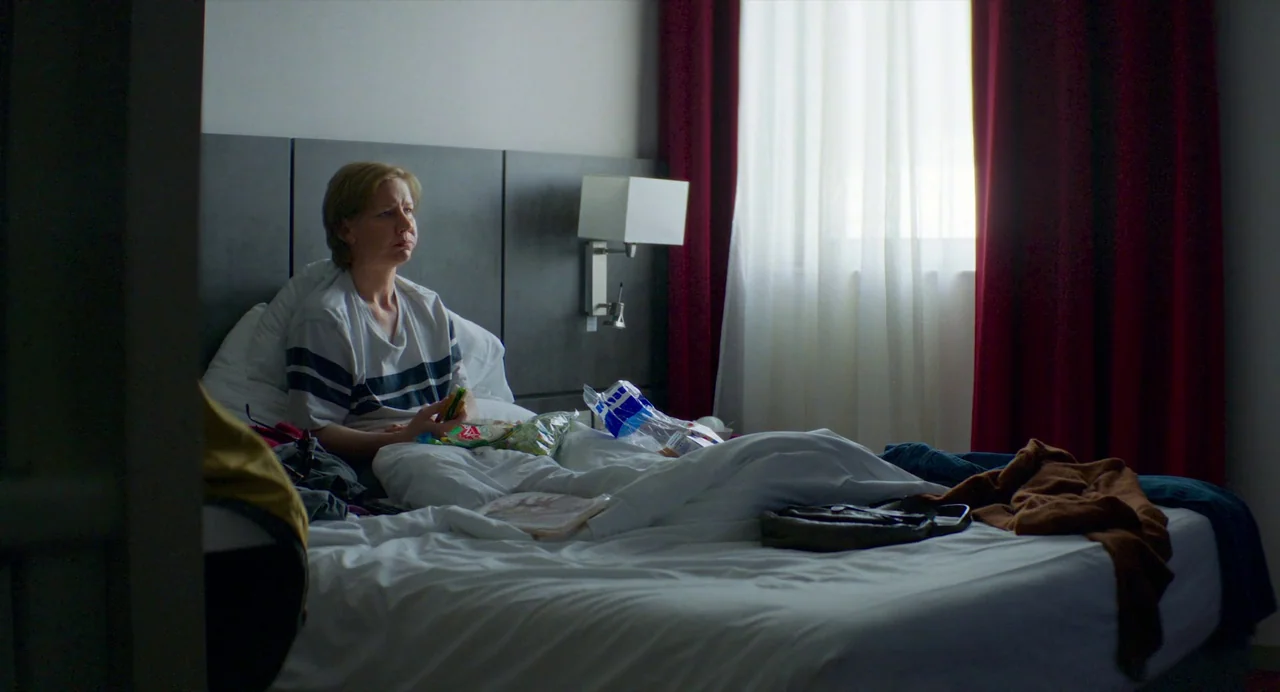 Anatomy of a Fall (2023) movie still: medium — A modern hotel room with dark wall panels and red curtains by a window. A woman sits in the bed among…