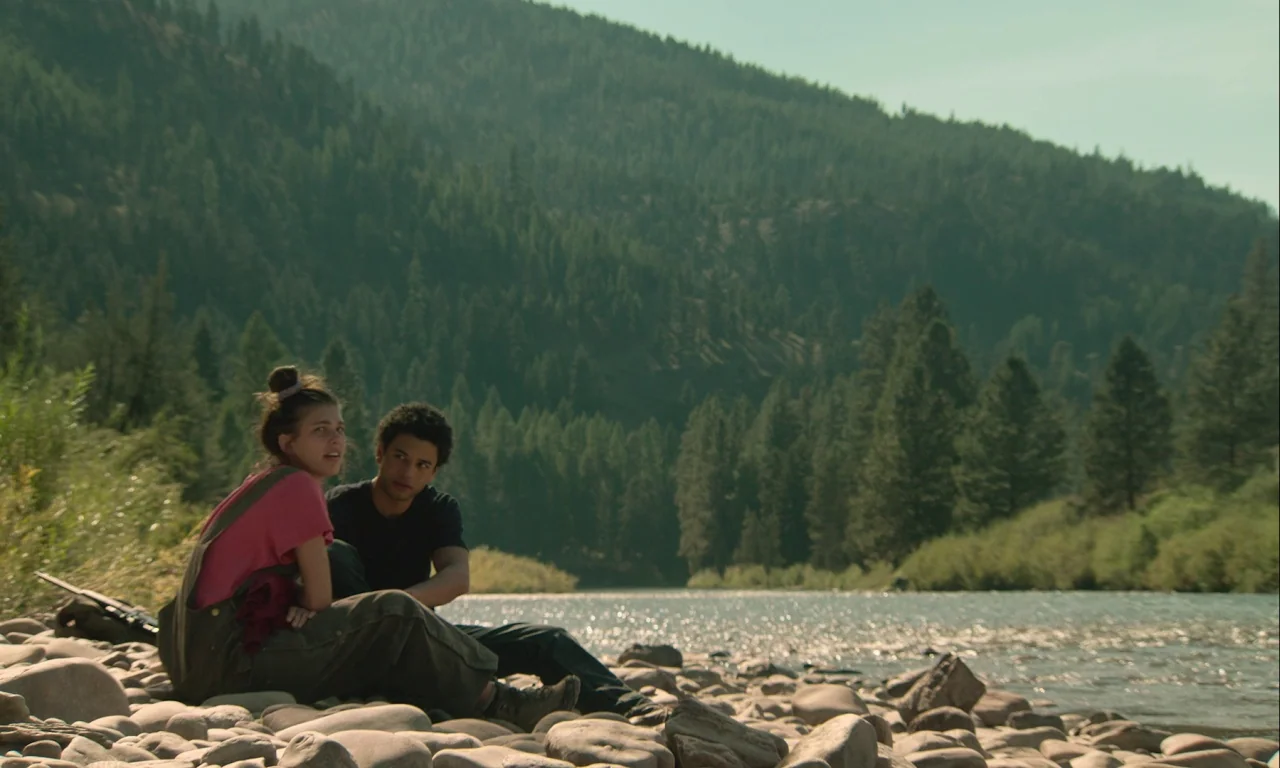 Mickey and the Bear (2019) movie still: wide — Two teenagers sit on a rocky riverbank in front of a dense pine forest. The pair rests on the stones…