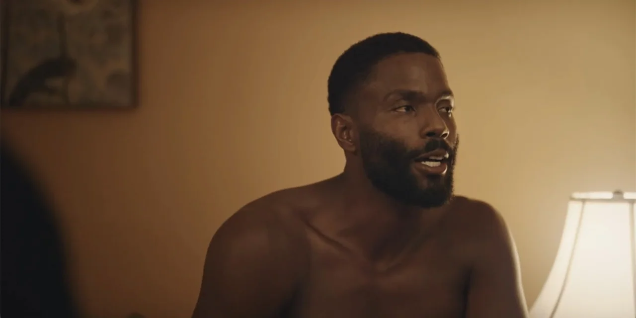 Equal Standard (2021) movie still: medium — A dimly lit bedroom with plain tan walls and a visible table lamp. A shirtless Black man in his thirties…