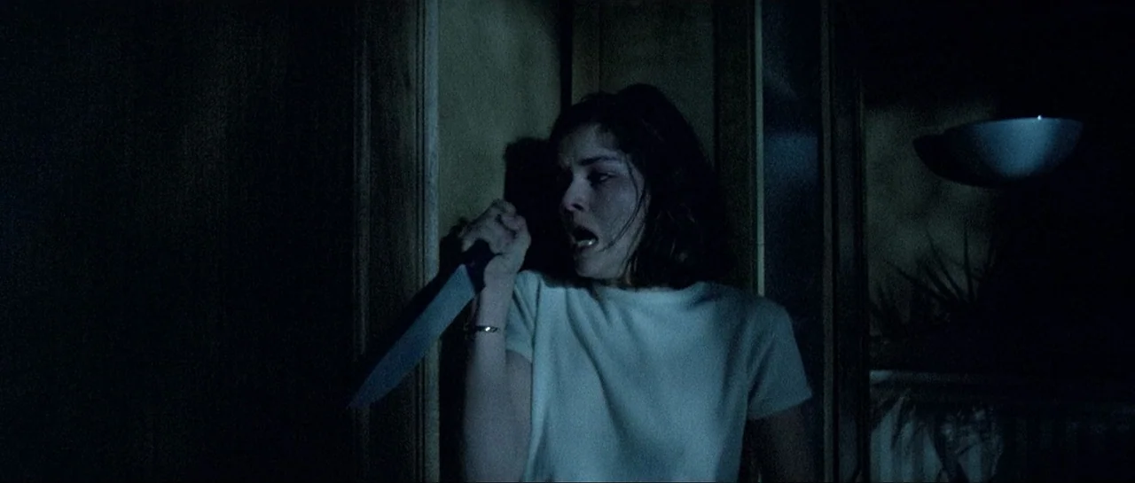 Opera (1987) movie still: medium — A dark, narrow residential hallway with wooden walls is cast in low-key lighting. A young woman stands against…