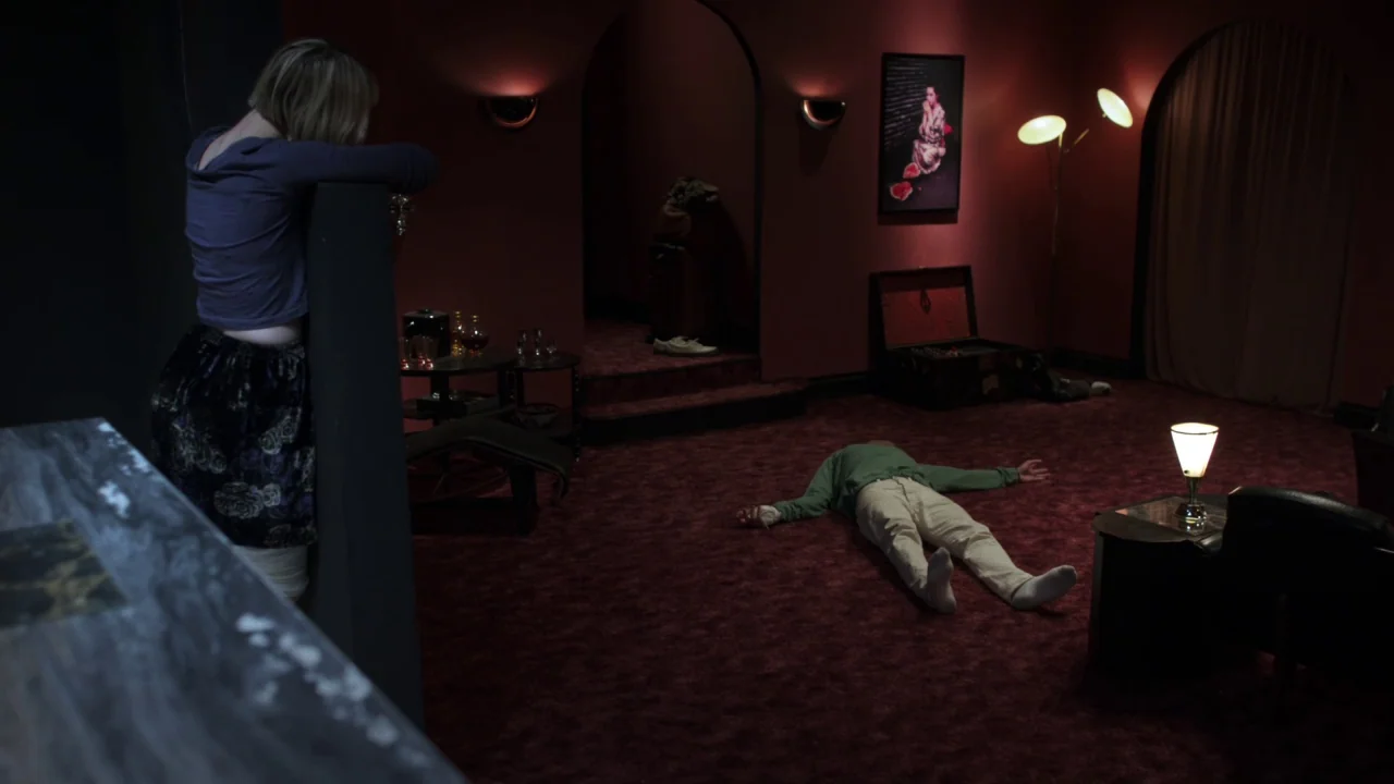 Piercing (2018) movie still: wide — A spacious room features deep red walls and carpeting with arched doorways and artwork. A man lies supine in the…