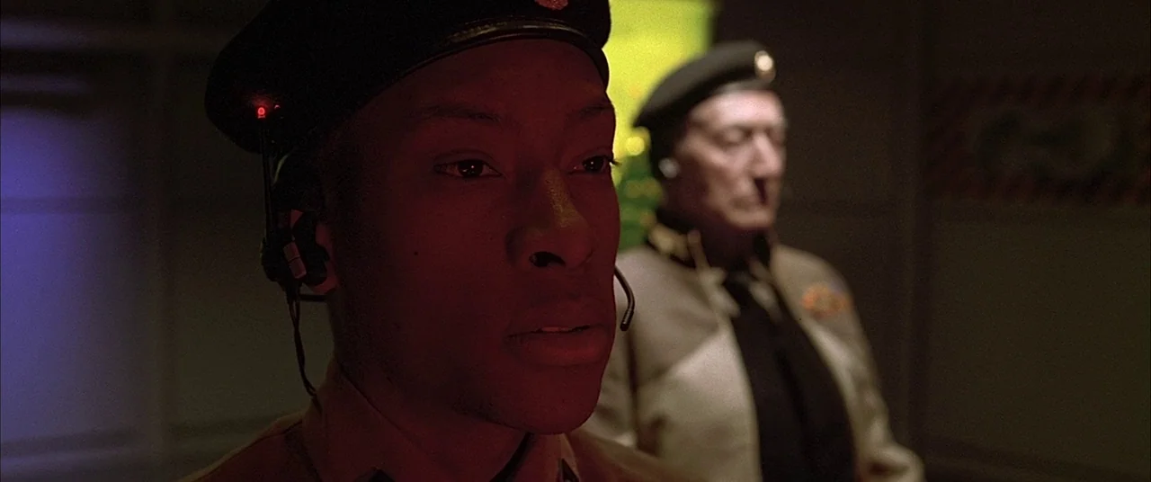 The Fifth Element (1997) movie still: medium — A dimly lit room features a younger man in a headset in the foreground. An older military officer…