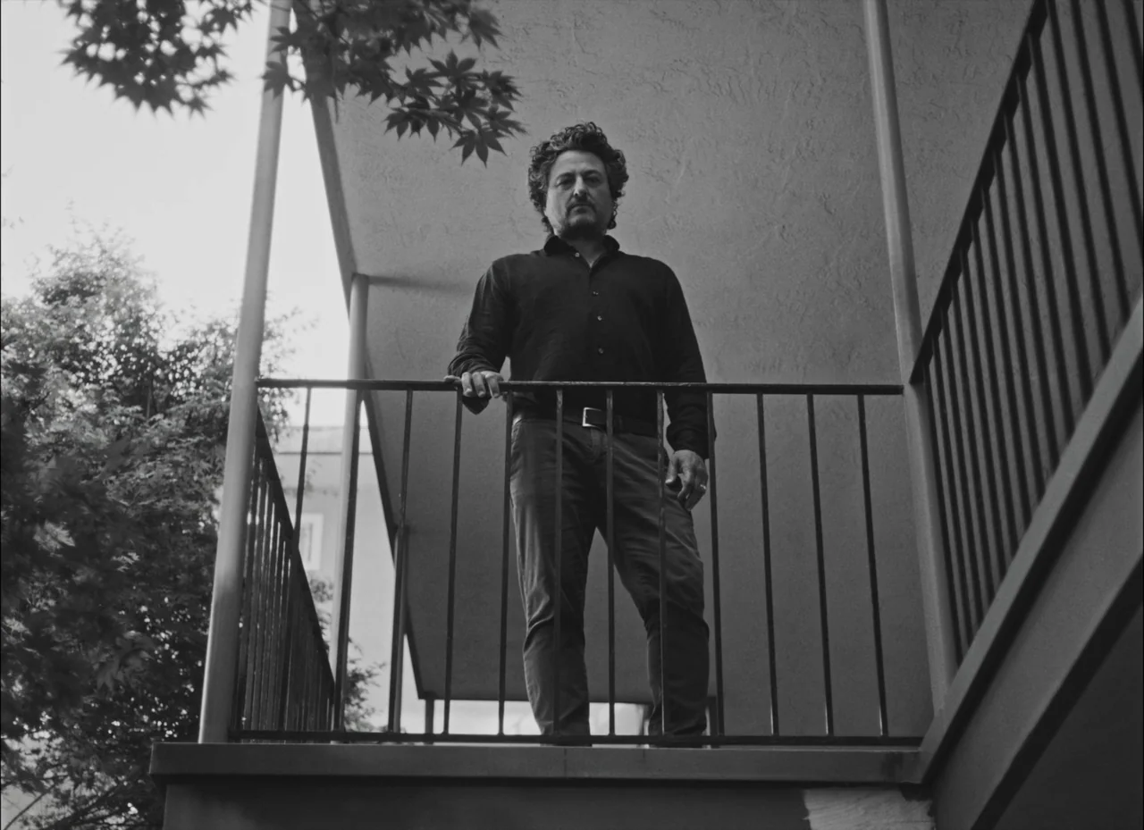 Fremont (2023) movie still: fullbody — An exterior residential building walkway with metal railings and concrete construction. The man stands on the…
