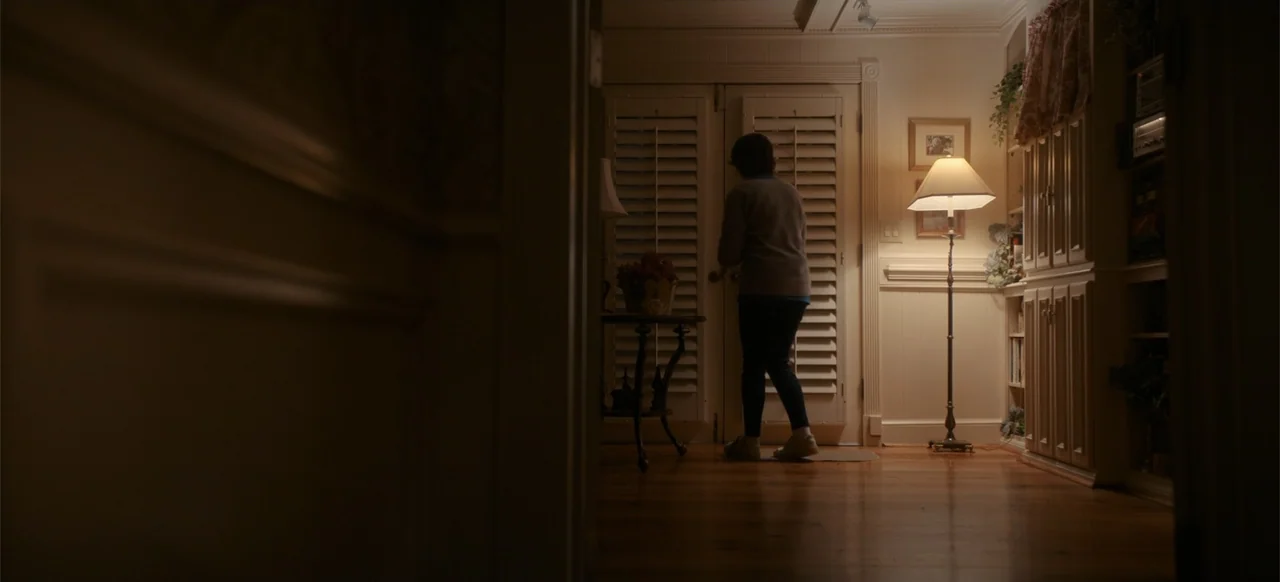 My Best Friend's Exorcism (2022) movie still: fullbody — A dimly lit room with hardwood floors and built-in shelving featuring closed shutter doors.…