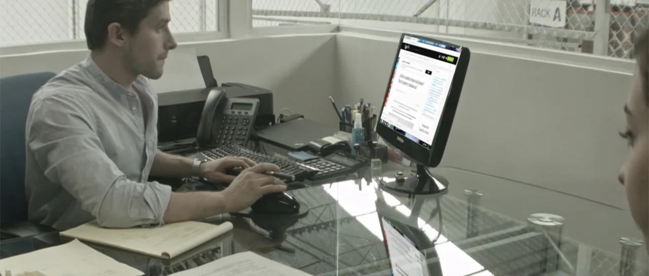Amor Mutante (2019) movie still: medium — An office interior features a glass-topped desk with a modern computer and office supplies. A man wearing…