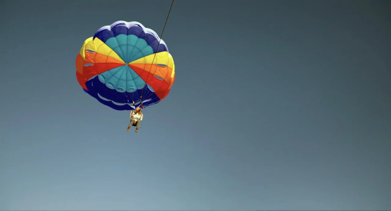 Blended (2014) movie still: wide — A clear sky provides a vast background for a person floating under a parachute. A brightly colored circular…