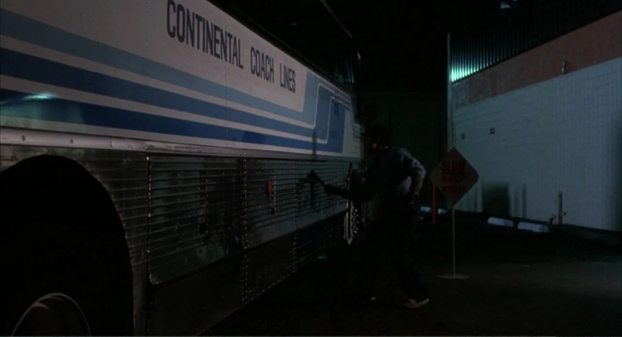 Murphy's Law (2009) movie still: wide — A night exterior shot of a large bus parked against a white tiled wall. A lone person stands beside the bus…