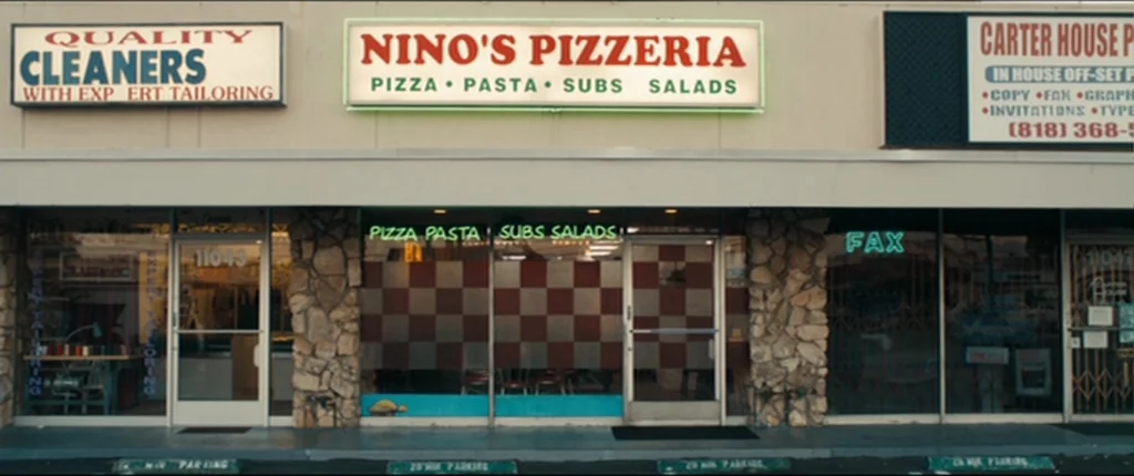 Drive (2011) movie still: wide — A strip mall at night featuring three storefronts with illuminated signs. Nino's Pizzeria occupies the central unit…
