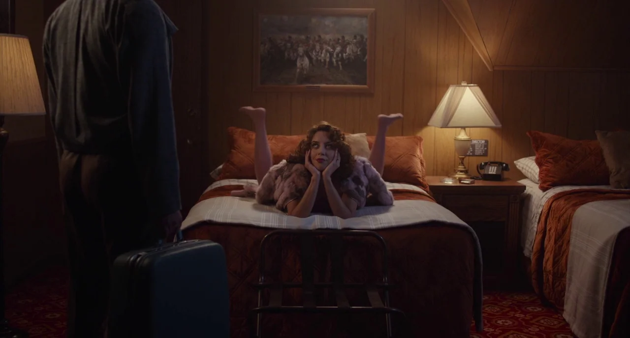 An Evening With Beverly Luff Linn (2018) movie still: medium — A 1970s style motel room features wood-paneled walls and orange quilted bedding. A…