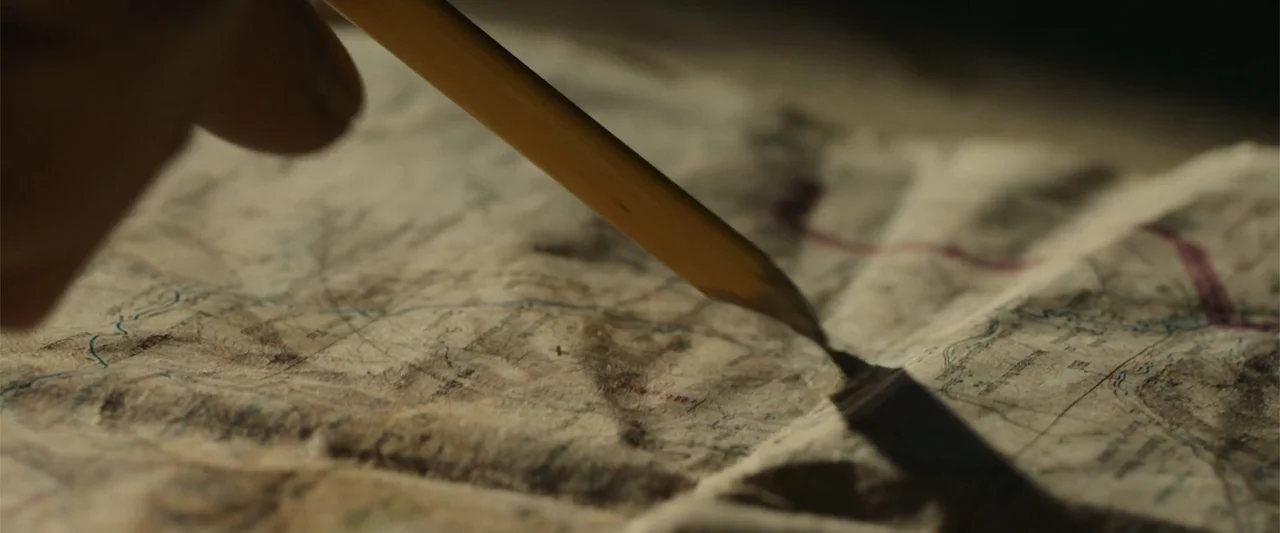 12 Strong (2018) movie still: closeup — An aged, creased topographical paper map lies flat on a dark surface. A yellow wooden pencil rests against…