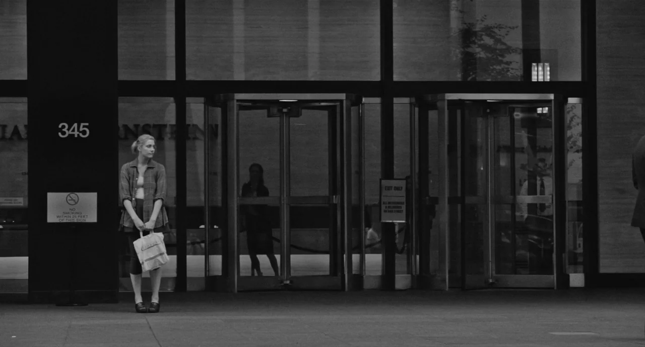 Frances Ha (2012) movie still: fullbody — A woman stands before the glass entrance of a large office building with revolving doors. She holds a…
