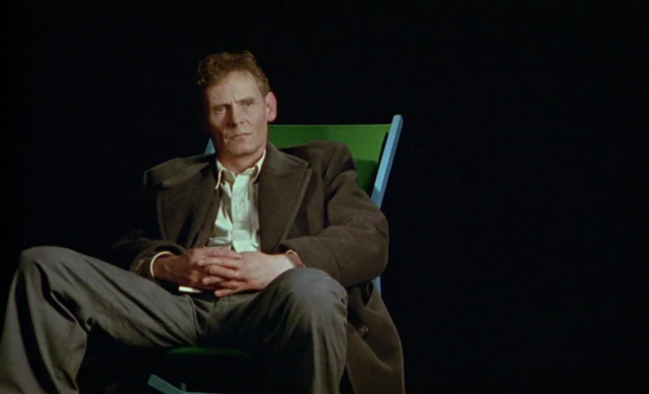 Wittgenstein (1993) movie still: medium — A man sits in a folding chair against a solid black backdrop. He holds his hands together while looking…
