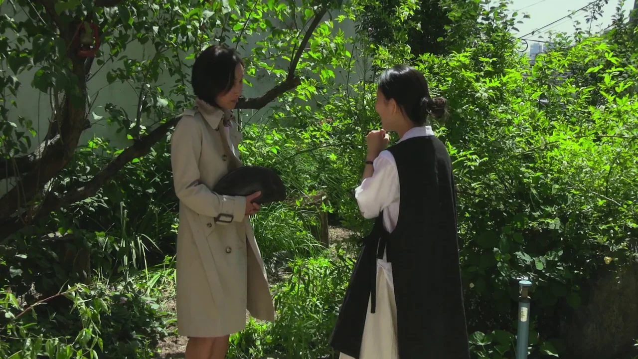 In Front Of Your Face (2021) movie still: fullbody — Two women stand in a sunlit area filled with dense green leafy bushes. One woman wears a trench…
