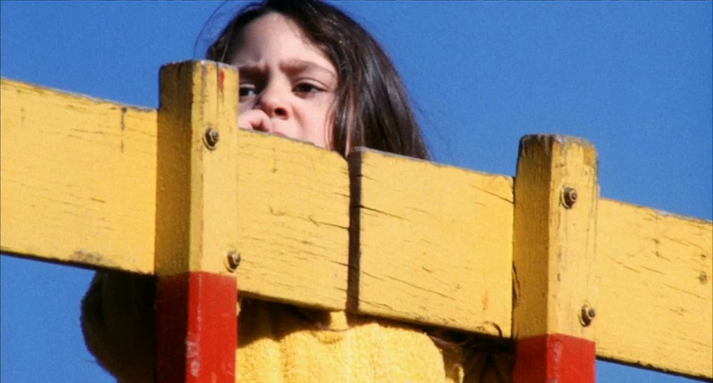A Woman Under the Influence (1974) movie still: closeup — A child peers over a bright yellow painted wooden structure against a blue sky. The wooden…