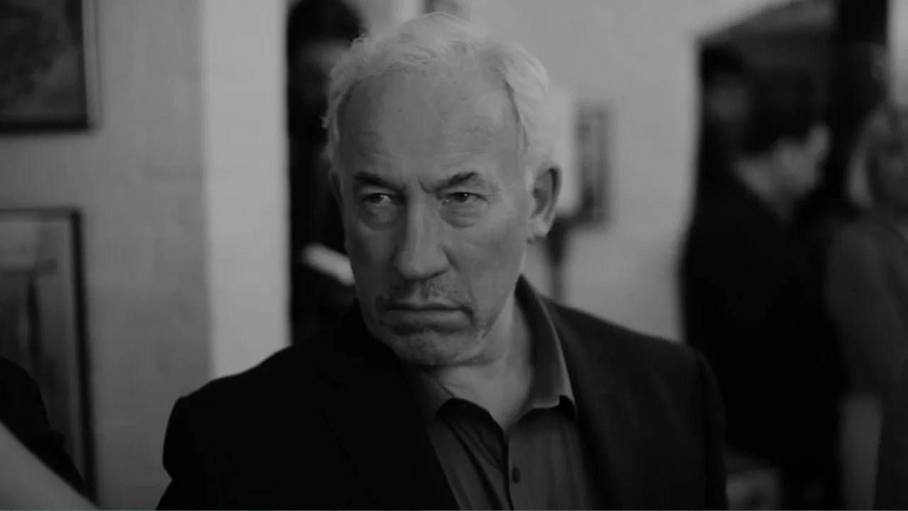 Creditors (2025) movie still: medium — A dimly lit indoor hallway with a white wall and a framed picture visible. An older man in a dark suit stands…