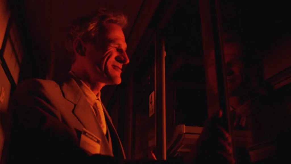 The Ugly Swans (2006) movie still: medium — A dark train car interior is illuminated solely by intense red cabin lights. A man in a suit looks…