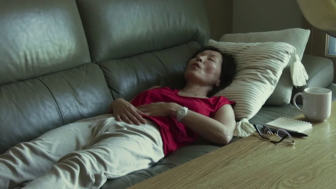 In Front Of Your Face (2021) movie still: medium — A woman rests on a dark green leather sofa in a residential space. A side table nearby holds a…