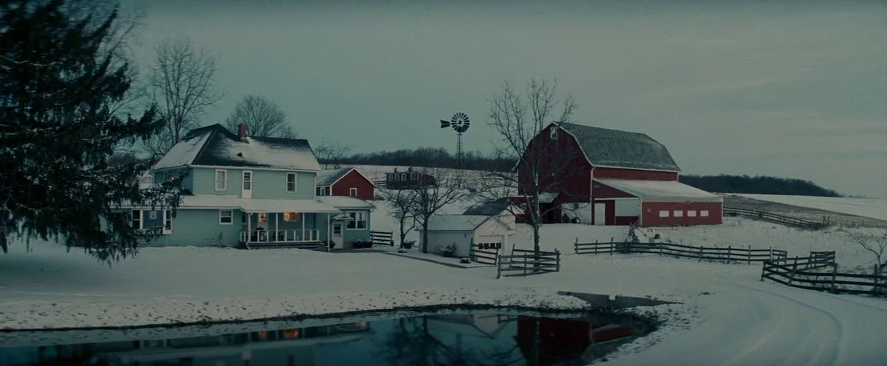 Michael Clayton (2007) movie still: establishing — A rural property with a light blue house and red barn sits in snow. A windmill stands between the…