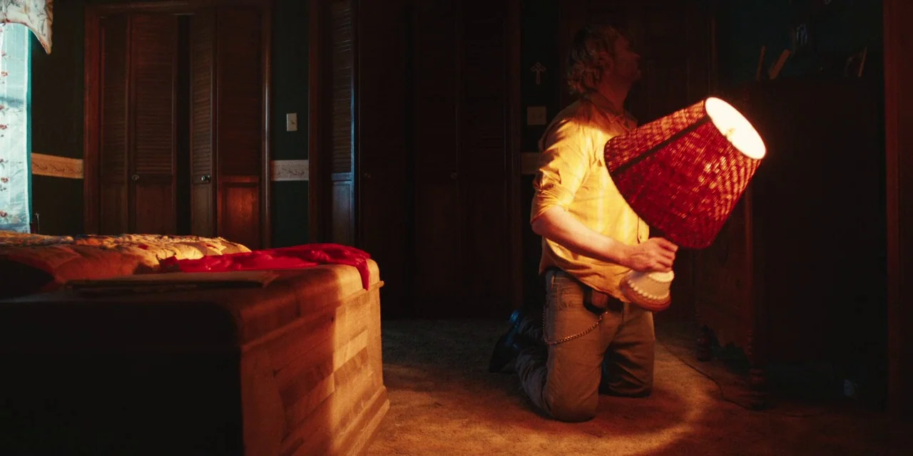 The Death of Dick Long (2019) movie still: medium — A dark, carpeted bedroom features a wooden trunk and closed louvered doors in the background. A…