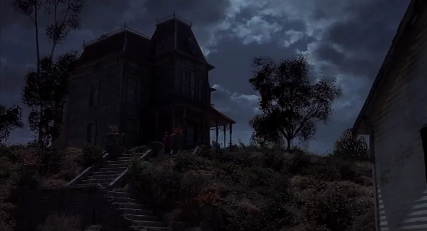 Psycho 2 (1983) movie still: wide — A dark Victorian mansion sits atop a grassy hill with stone stairs leading up. Clouds drift across a night sky…