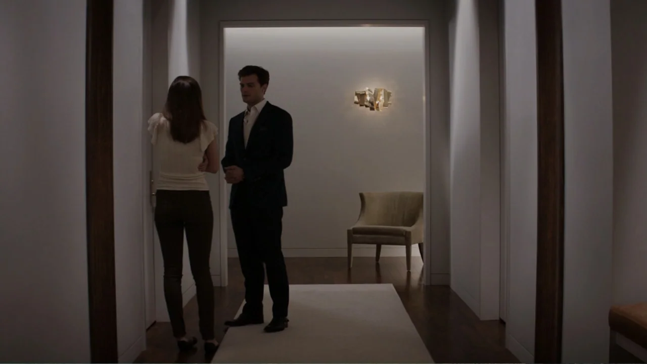 Fifty Shades of Grey (2015) movie still: fullbody — A minimalist corridor features white walls, dark flooring, and a single sculptural gold lamp. A…