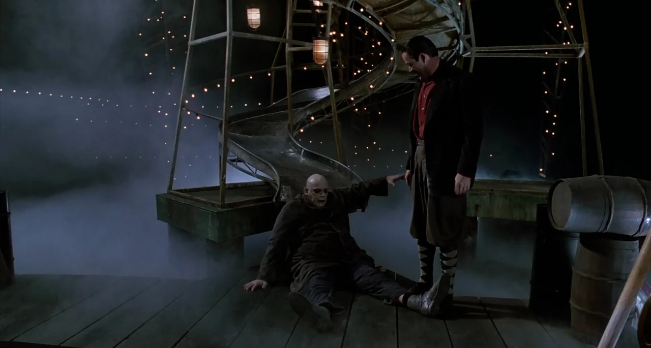 The Addams Family (1991) movie still: fullbody — A wooden pier at night is covered in heavy fog with a spiral slide structure. Two men stand…