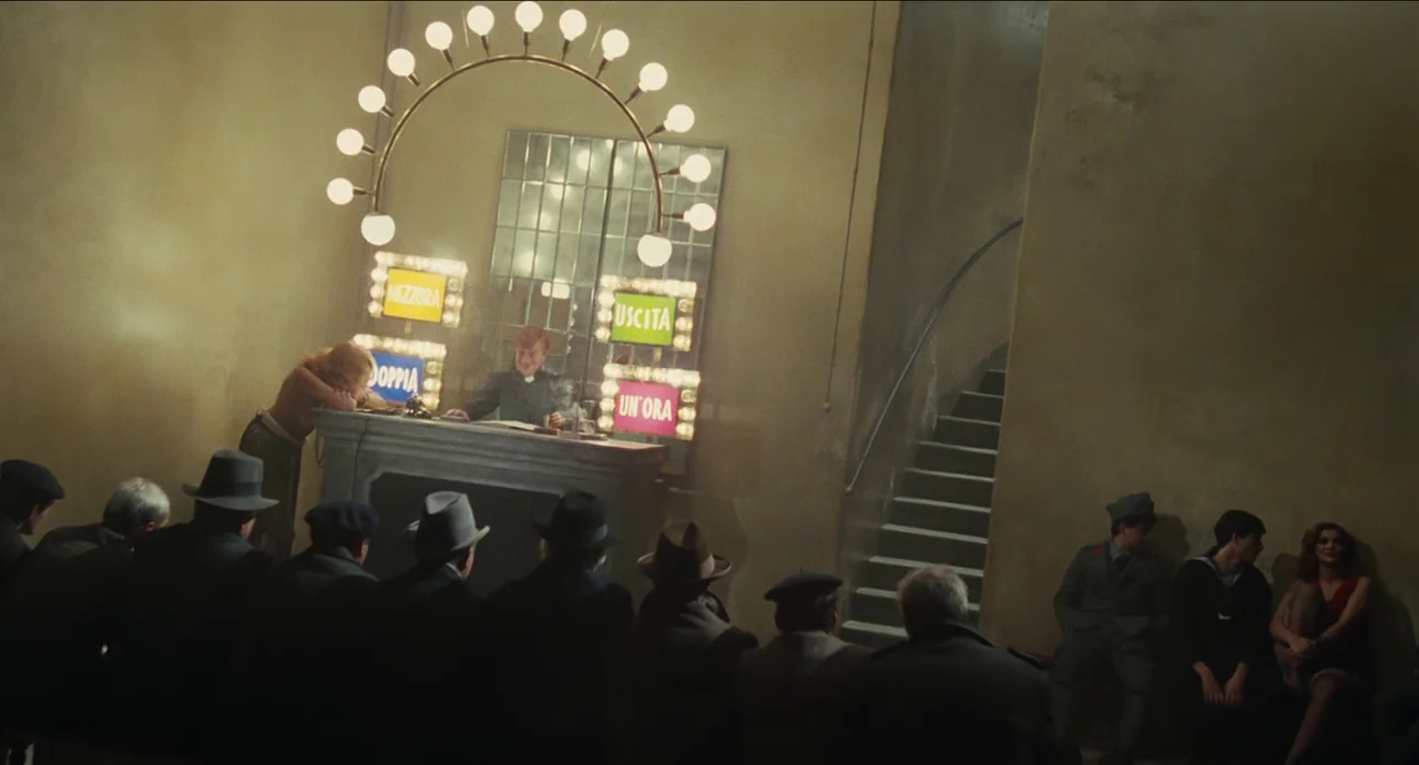 City of Women (1980) movie still: wide — A spacious, dimly lit lobby features a central reception desk illuminated by glowing signs. A group of men…