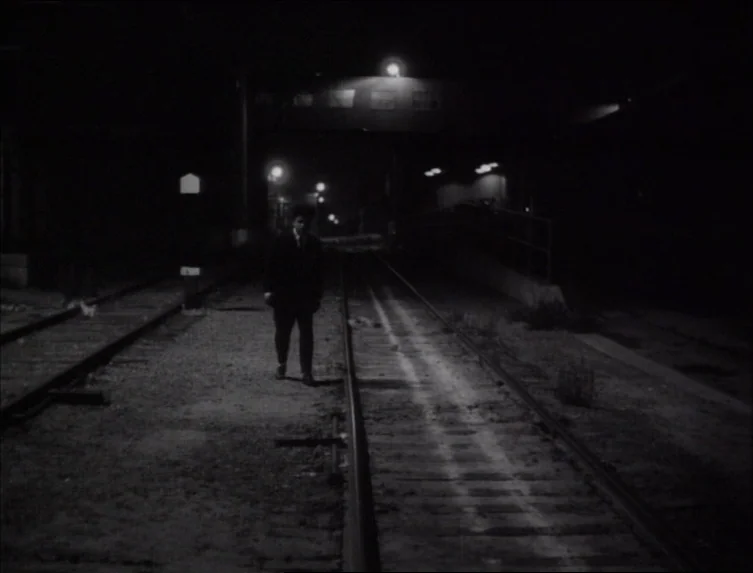 Eraserhead (1977) movie still: wide — A man stands between two sets of train tracks under the glow of street lamps. The surrounding area is dark…