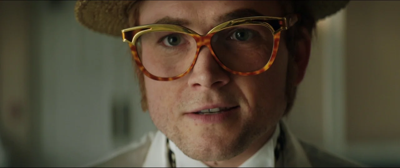 Rocketman (2019) movie still: closeup — An interior space features a man wearing a straw hat and distinctive spectacles. He looks directly at the…