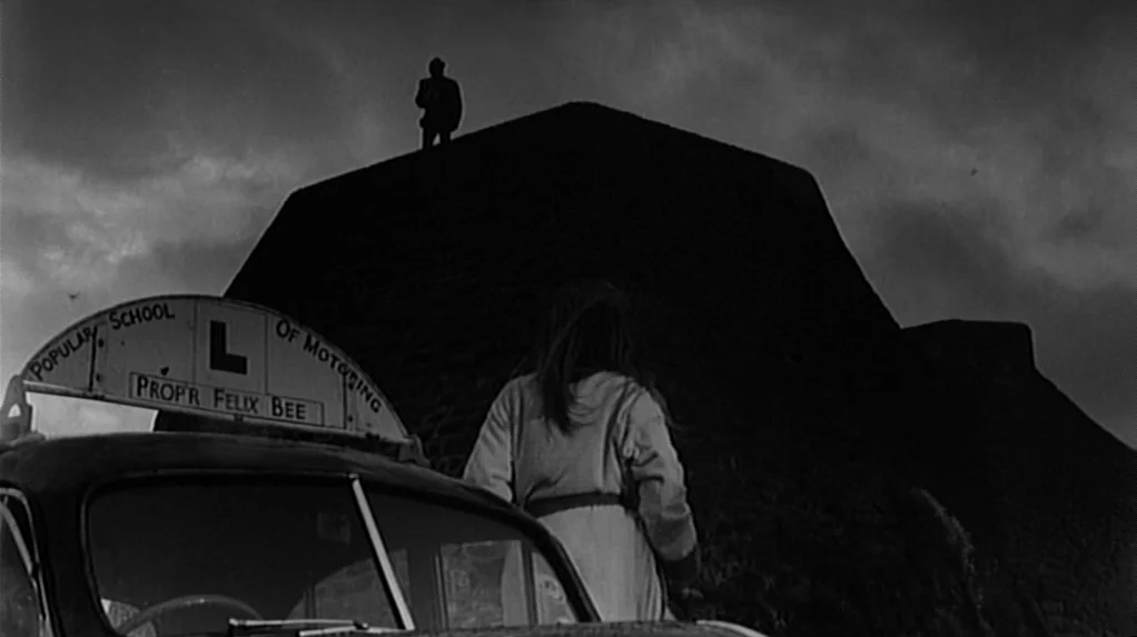 Cul-de-sac (1966) movie still: wide — A woman looks toward a silhouetted figure standing atop a large, dark stone mound. A learner driver car with a…